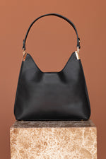 Load image into Gallery viewer, Back view of black vegan handbag with gold hardware sitting on a stone plinth.

